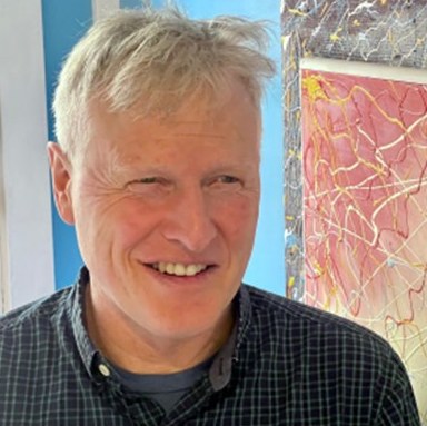 Paul Reynolds, smiling, in a checked shirt, with a colorful painting behind him